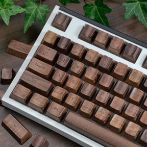 Puede incluir: Primer plano de un teclado mecánico con teclas de madera. Las teclas son de color marrón cálido, con letras blancas. El teclado tiene un marco blanco y una barra espaciadora marrón oscuro. Algunas teclas están sueltas.