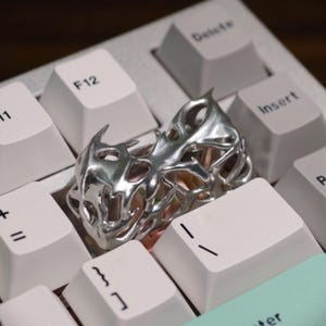 May include: A silver-colored ring with an intricate, openwork design, resembling an abstract organic form. The ring is placed on a white keyboard, with the keys visible in the background. The ring's design features sharp, pointed elements.