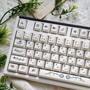 May include: A white mechanical keyboard with keycaps featuring floral designs in shades of blue, red, and green. The keyboard has a standard QWERTY layout with a black frame and is surrounded by greenery.
