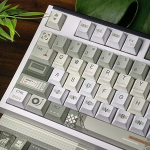 May include: A white mechanical keyboard with custom keycaps. The keycaps are a mix of white, gray, and green with various icons and text. The keyboard has a white frame and is partially visible.
