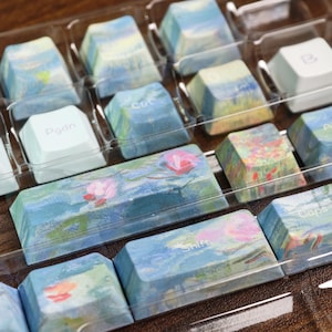 May include: A collection of mechanical keyboard keycaps featuring an impressionist-style painting of water lilies in shades of blue, green, and pink. The set includes standard keys with white text such as Ctrl, Pgdn, Shift, and Caps, housed in a clear plastic tray.