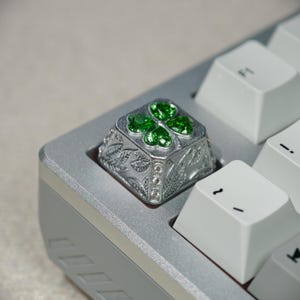 May include: A silver keyboard keycap featuring a four-leaf clover design. The keycap has green jewel-like accents on the clover and small clear gems around the sides. The keycap is set on a silver keyboard.