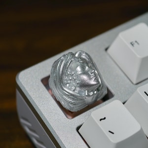 Op de afbeelding: Een zilverkleurige keycap met een gedetailleerd gebeeldhouwd gezicht van een vrouw met vloeiend haar. De keycap is geïnstalleerd op een lichtgrijs mechanisch toetsenbord, naast witte toetsen met zwarte letters.