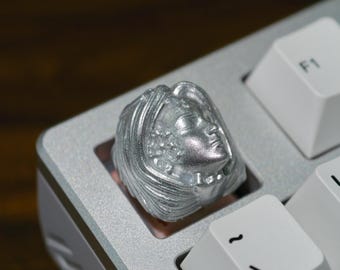 May include: A silver-colored keycap featuring a detailed sculpted face of a woman with flowing hair. The keycap is installed on a light gray mechanical keyboard, next to white keys with black lettering.