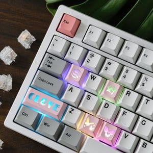 May include: White mechanical keyboard with custom keycaps. The keyboard has a pink Escape key and a pink keycap that says "CLOVE" in blue. Other keycaps have symbols and are illuminated with various colors.