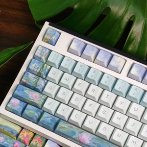 May include: A custom mechanical keyboard with a white frame and light blue keycaps. Some keycaps feature a watercolor design with blue, pink, and green hues. The keyboard has a standard QWERTY layout.