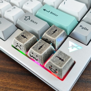 May include: Close-up of a mechanical keyboard with custom keycaps. The keycaps feature a gray, textured design with lightning bolt symbols and various facial expressions. Other keys are white and a teal Enter key is visible.