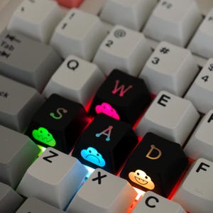 May include: A close-up of a mechanical keyboard with white and black keycaps. The black keycaps spell out "SAD" and "WA" with cloud graphics. The keys are backlit with various colours, including pink, green, blue, and orange.