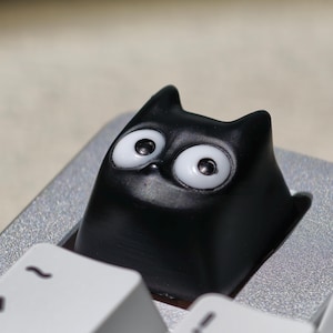 Può includere: Un copritasto nero a forma di gatto con grandi occhi bianchi e finitura lucida. Il copritasto è installato su una tastiera argentata, con un tasto bianco visibile sotto. Le orecchie del gatto sono a punta e gli occhi sono grandi ed espressivi.