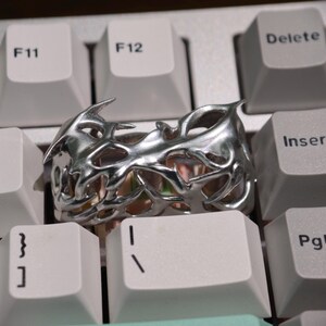 May include: A silver-colored ring with an intricate, openwork design featuring sharp, angular shapes. The ring is set against a white keyboard, highlighting its metallic sheen and unique style. The ring's design suggests a modern or fantasy theme.