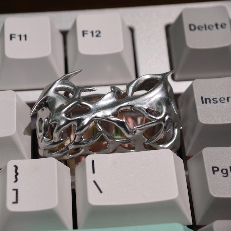 May include: A silver-colored ring with an intricate, openwork design featuring sharp, angular shapes. The ring is set against a white keyboard, highlighting its metallic sheen and unique style. The ring's design suggests a modern or fantasy theme.