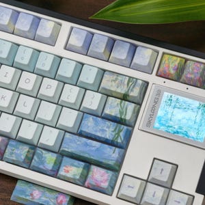 May include: A mechanical keyboard featuring a white frame and a mix of light blue, pale green, and impressionist-style floral patterned keycaps. A small rectangular screen on the right side displays a water lily pond image with the text SKYLOONG 3.0.