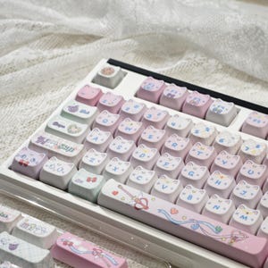 May include: A white mechanical keyboard with custom keycaps. The keycaps are primarily pink and white, featuring cat faces and other cute designs. Some keycaps have text and symbols. The keyboard is on a white surface.