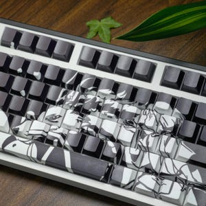 May include: A mechanical keyboard with black and white keycaps featuring an anime-style design. The keyboard has a white frame and a variety of keycap designs. The keycaps are a mix of solid black and black and white patterned designs.