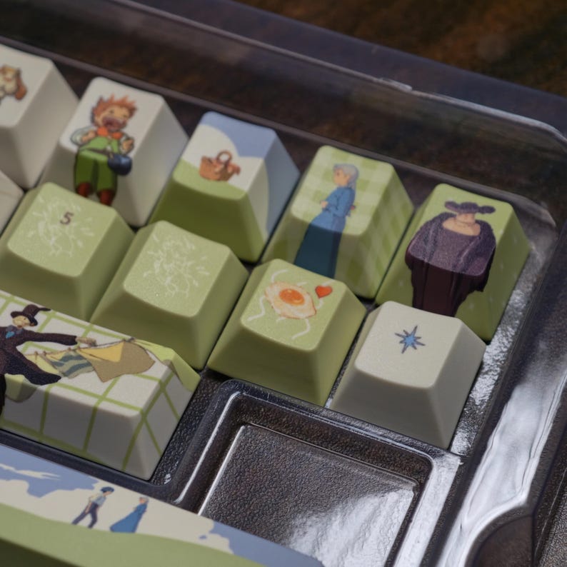 May include: A set of custom keyboard keycaps featuring illustrations of characters and scenes. The keycaps have a variety of designs, including a person, a basket, and a fried egg. The keycaps are in a clear plastic container.