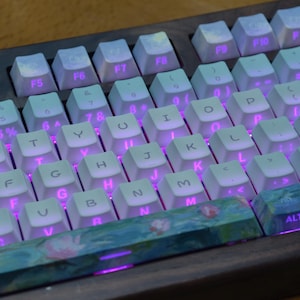 May include: A mechanical keyboard featuring white keycaps with black lettering and vibrant purple backlighting. The spacebar and bottom row keys display a colorful floral pattern. The keyboard is set within a dark wooden frame.