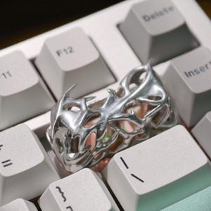 May include: A silver-colored ring with an intricate, openwork design featuring spiky, flame-like elements. The ring is displayed on a white keyboard, highlighting its metallic sheen and detailed craftsmanship. The ring's design suggests a gothic or fantasy theme.
