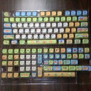 Puede incluir: Un conjunto de teclas de teclado coloridas con un dise&ntilde;o caprichoso. Las teclas presentan una variedad de colores, incluyendo verde, azul, amarillo y blanco, con ilustraciones de dibujos animados. Las teclas est&aacute;n dispuestas en una bandeja de pl&aacute;stico transparente.