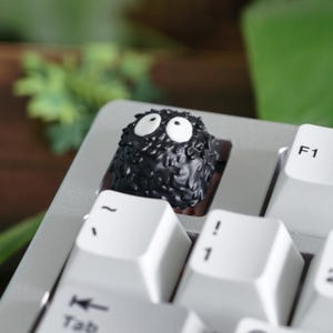 Op de afbeelding: Een zwarte, getextureerde artisan keycap met grote witte ogen, die lijkt op een roetelf, geïnstalleerd op een zilverkleurig toetsenbord. De keycap wordt omringd door witte toetsen met zwarte letters, inclusief de F1-toets.