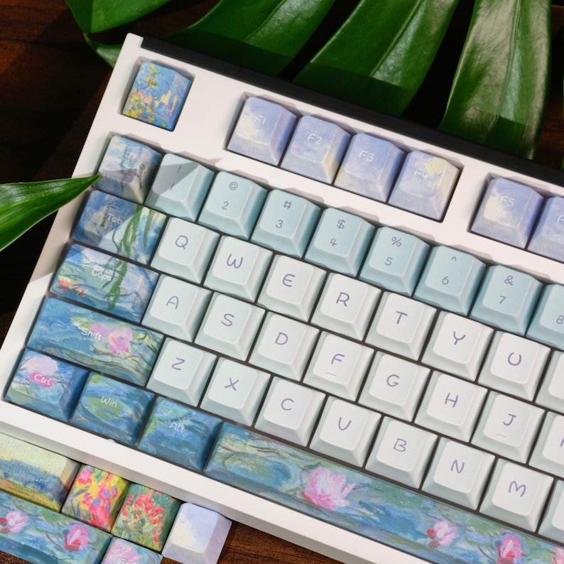 May include: A mechanical keyboard featuring a white frame and a custom keycap set. The keys display a light blue and white color scheme, with specific keys decorated in a colorful impressionist floral pattern featuring pink and green tones.