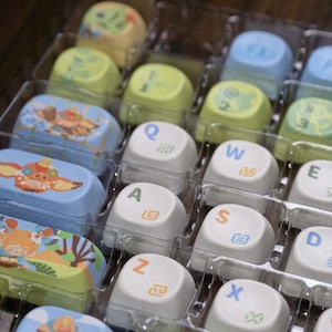 Puede incluir: Una colecci&oacute;n de teclas de teclado personalizadas en una bandeja de pl&aacute;stico transparente. Las teclas presentan varios dise&ntilde;os y colores, incluyendo blanco, azul, verde y amarillo. Algunas teclas tienen letras y s&iacute;mbolos, otras ilustraciones de dibujos animados.