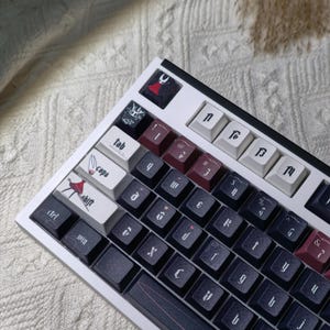 Può includere: Una tastiera meccanica bianca con tasti neri e bordeaux. La tastiera presenta tasti personalizzati con design e simboli unici. La tastiera è su uno sfondo bianco strutturato.