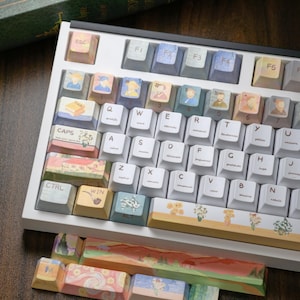 Peut inclure: Un clavier mécanique blanc avec des touches personnalisées. Les touches présentent des illustrations à l'aquarelle de portraits, de paysages et de motifs floraux. Le clavier a un cadre blanc et une variété de couleurs de touches, dont le bleu, le jaune et le vert.