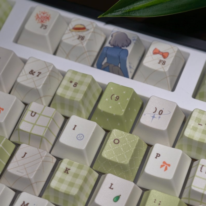 May include: Close-up of a keyboard with custom keycaps. The keycaps feature a mix of white and green patterns, including gingham and floral designs. Some keycaps have illustrations of a character in a blue dress, a bow, and a hat. The keyboard has the letters and numbers.