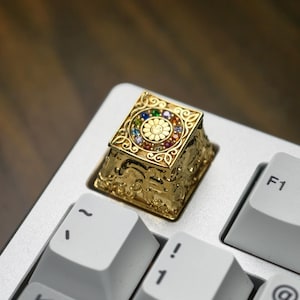 May include: A gold-colored, ornate keyboard keycap with a square top and intricate floral and scroll designs. The top features a circular design with colorful gemstones and a central flower. The keycap is set on a silver keyboard.