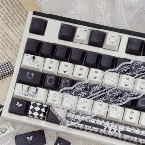 May include: A white and black mechanical keyboard with custom keycaps. The keycaps feature various designs, including butterflies, hearts, and geometric patterns. A decorative black and white lace ribbon is placed across the keyboard.