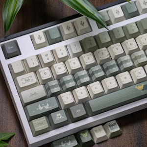 Puede incluir: Un teclado mecánico personalizado con marco blanco y teclas en tonos verdes y crema. Las teclas presentan diseños decorativos, incluyendo motivos florales, mariposas y encaje. El teclado tiene una distribución estándar con teclas de función.