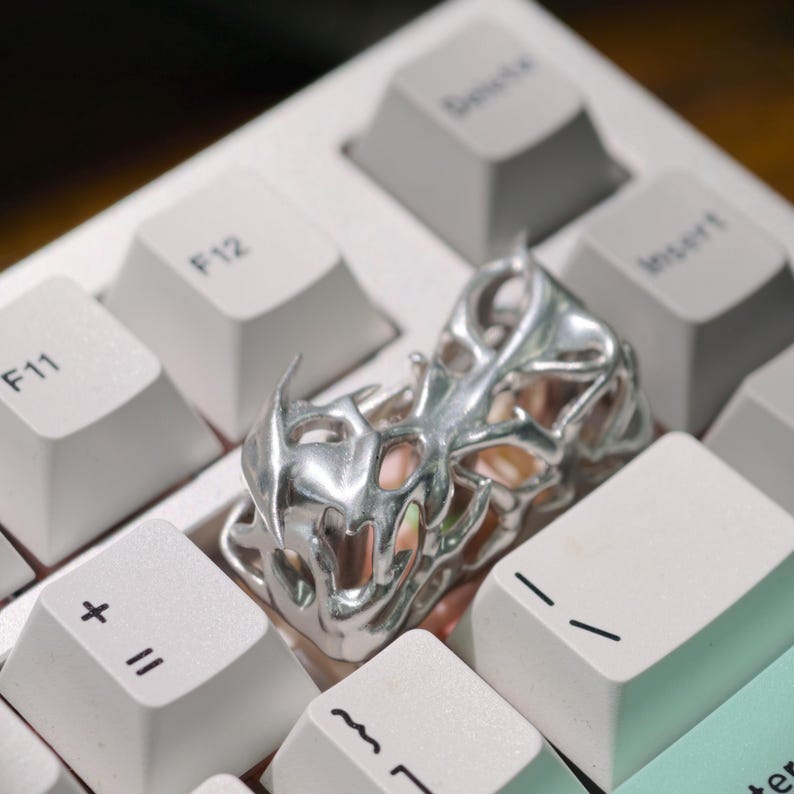 May include: A silver-colored, abstract ring with an open, intricate design rests on a white keyboard. The ring's form is organic, with multiple openings and a textured surface. The keyboard keys are white with gray lettering.