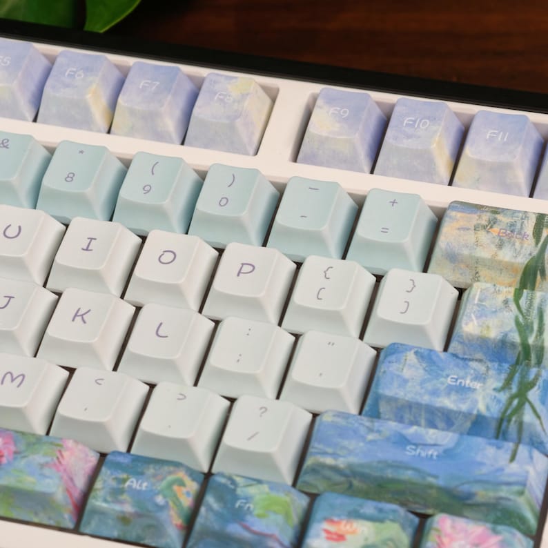 May include: A mechanical keyboard featuring a mix of solid white keycaps with dark lettering and custom keycaps decorated with blue, green, and pink impressionist-style floral patterns. The layout includes function keys, numbers, and letters.