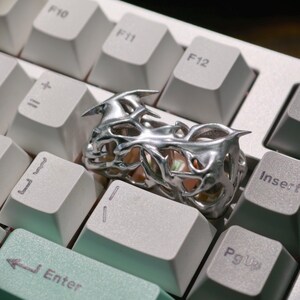 May include: A silver-colored ring with an intricate, openwork design featuring sharp, pointed elements. The ring is placed on a white keyboard, highlighting its metallic sheen and unique shape. The ring's design suggests a gothic or fantasy theme.
