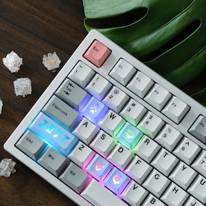 May include: A white mechanical keyboard with illuminated keys. The keyboard has a pink Esc key and several custom keycaps with unique designs. Additional clear key switches are scattered around the keyboard on a wooden surface.
