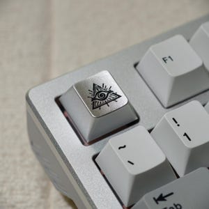 May include: Close-up of a silver mechanical keyboard with white keycaps. The F1 key is visible, along with other keys. The keycap in focus has a black graphic of an eye inside a triangle.