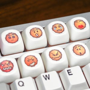 May include: White keyboard keys with various emoji faces. The emoji faces include expressions of anger, sadness, and joy. The keys are arranged in rows and columns, with the letters Q, W, and E visible on the bottom row.