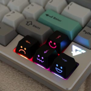 以下が含まれることがあります： カスタムキーキャップを備えたメカニカルキーボードのクローズアップ。キーキャップには、さまざまな絵文字の顔と稲妻のデザインが施されています。キーボードは白とグレーの配色で、ターコイズ色のEnterキーが付いています。