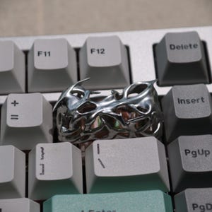 May include: A silver-colored ring with an intricate, openwork design, resting on a computer keyboard. The ring's surface reflects light, highlighting its detailed, organic pattern. The keyboard keys are light gray and dark gray.