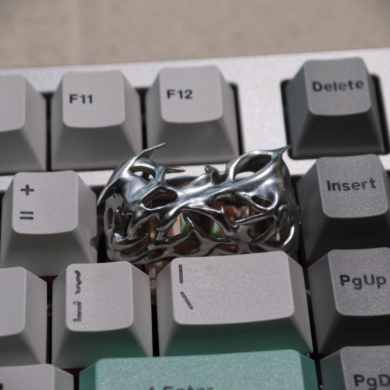 May include: A silver-colored ring with an intricate, openwork design, resting on a computer keyboard. The ring's surface reflects light, highlighting its detailed, organic pattern. The keyboard keys are light gray and dark gray.