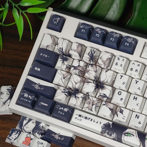 Może przedstawiać: Biała klawiatura mechaniczna z niestandardowymi nakładkami na klawisze. Nakładki na klawisze mają kwiatowy wzór w kolorze białym i szarym, z granatowymi klawiszami akcentującymi. Niektóre klawisze mają japońskie znaki. Klawiatura jest otoczona zielenią.