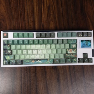 May include: A white mechanical keyboard with green and gray keycaps. The keycaps feature various illustrations, including forest scenes and characters. The keyboard has a small screen on the right side and a white frame.