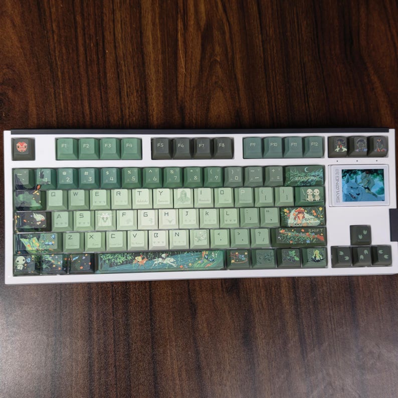 May include: A white mechanical keyboard with green and gray keycaps. The keycaps feature various illustrations, including forest scenes and characters. The keyboard has a small screen on the right side and a white frame.