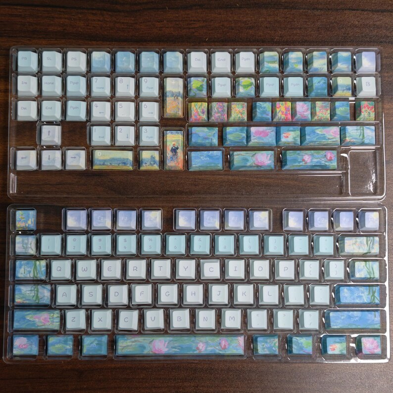 May include: A set of mechanical keyboard keycaps arranged in two clear plastic trays. The keycaps feature a mix of solid light blue and white keys alongside keys printed with impressionist-style floral and water lily landscape paintings.