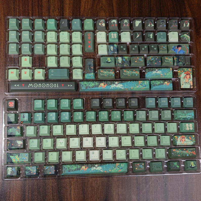 May include: A set of green and white keycaps for a mechanical keyboard, featuring custom artwork and the word "MONONOKE". The keycaps are arranged in a clear plastic tray, ready for installation. The design includes illustrations of characters and scenes.