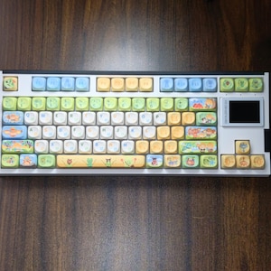Puede incluir: Un teclado mec&aacute;nico blanco con teclas coloridas. Las teclas presentan varias ilustraciones y dise&ntilde;os, incluyendo criaturas marinas, plantas y personajes de dibujos animados. El teclado tiene una peque&ntilde;a pantalla en el lado derecho y est&aacute; sobre una superficie de madera.