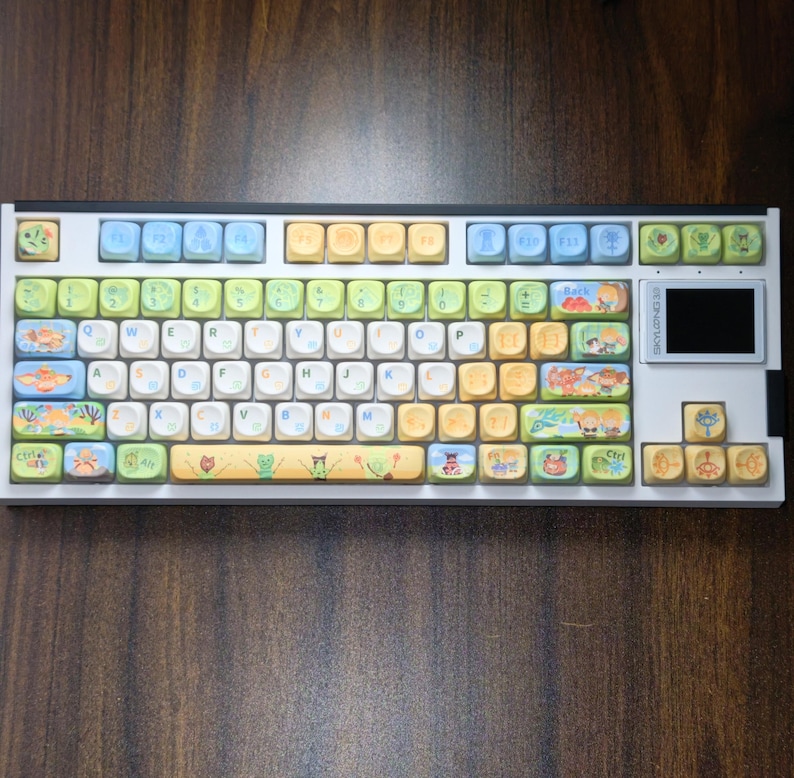 Puede incluir: Un teclado mec&aacute;nico blanco con teclas coloridas. Las teclas presentan varias ilustraciones y dise&ntilde;os, incluyendo criaturas marinas, plantas y personajes de dibujos animados. El teclado tiene una peque&ntilde;a pantalla en el lado derecho y est&aacute; sobre una superficie de madera.