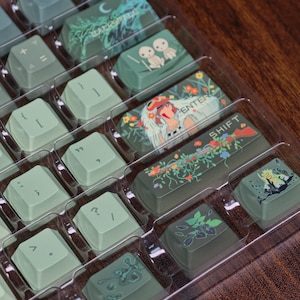 May include: A set of custom keyboard keycaps in a clear plastic tray. The keycaps are various shades of green and feature unique designs, including floral patterns and anime-style illustrations. Some keycaps have text like "ENTER" and "SHIFT".