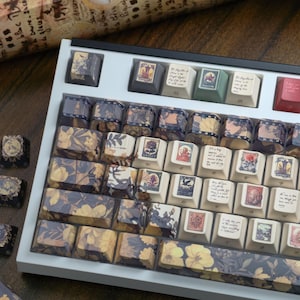 May include: A custom keyboard with keycaps featuring floral and vintage stamp designs. The keycaps have a variety of colors, including cream, yellow, blue, and red. The keyboard is set in a white frame.