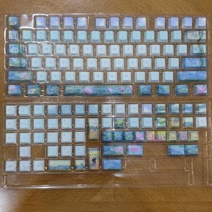 May include: A set of mechanical keyboard keycaps arranged in two clear plastic trays. The keycaps feature a white base with printed blue, green, and pink floral and landscape designs, including water lilies and figures in a field, with white letters.